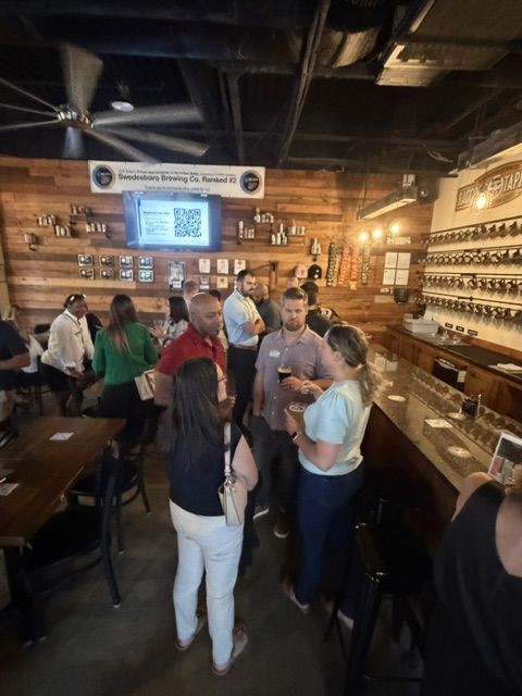 Attendees chatting along the bar while a Work The Room QR code shows on the TV at the Raccoon Taproom