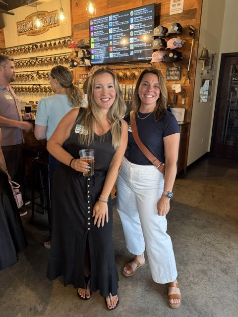 Two attendees smiling together in front of the Raccoon Taproom tap list and hat wall