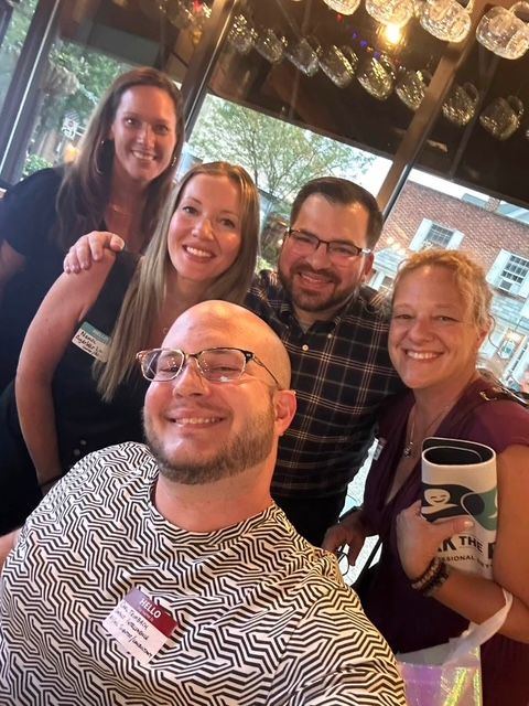 Five attendees with name tags squeezed into a smiling group selfie by a window at a Work The Room event